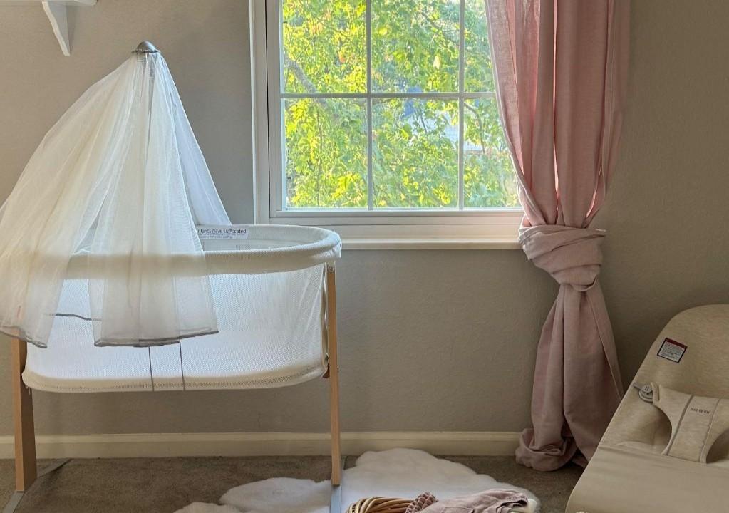 A serene nursery with a white bassinet under a net canopy by a sunlit window, pink curtains, and a cream-colored baby seat on the floor.