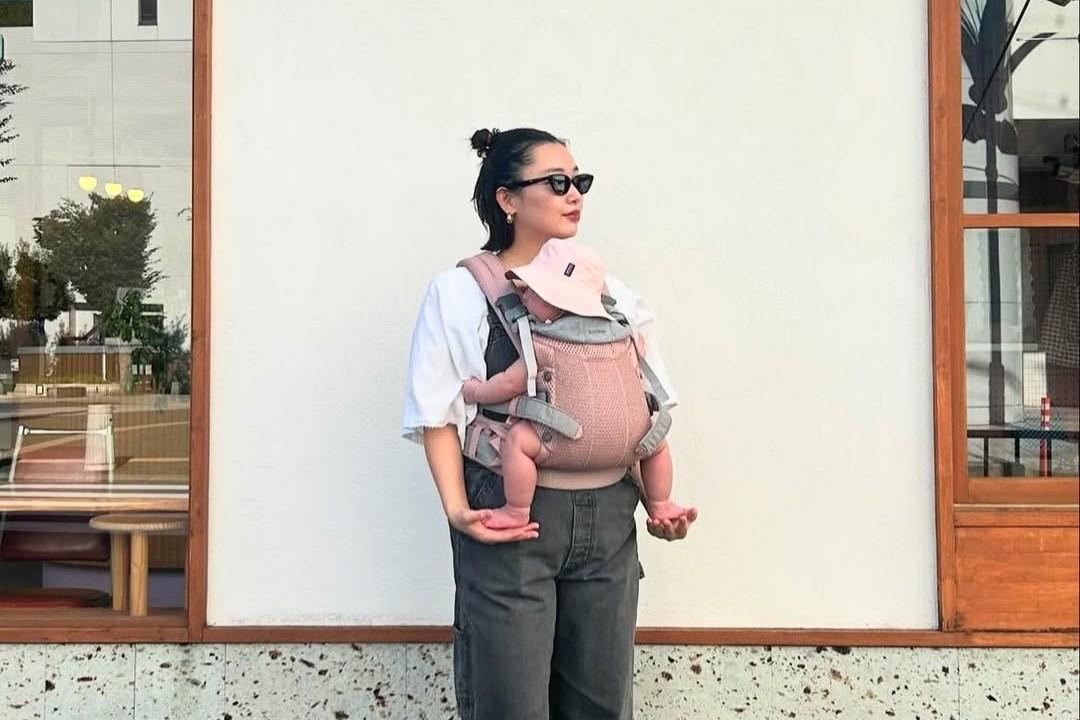 Woman wearing sunglasses holds a baby in a front carrier, standing against a white wall with a window.