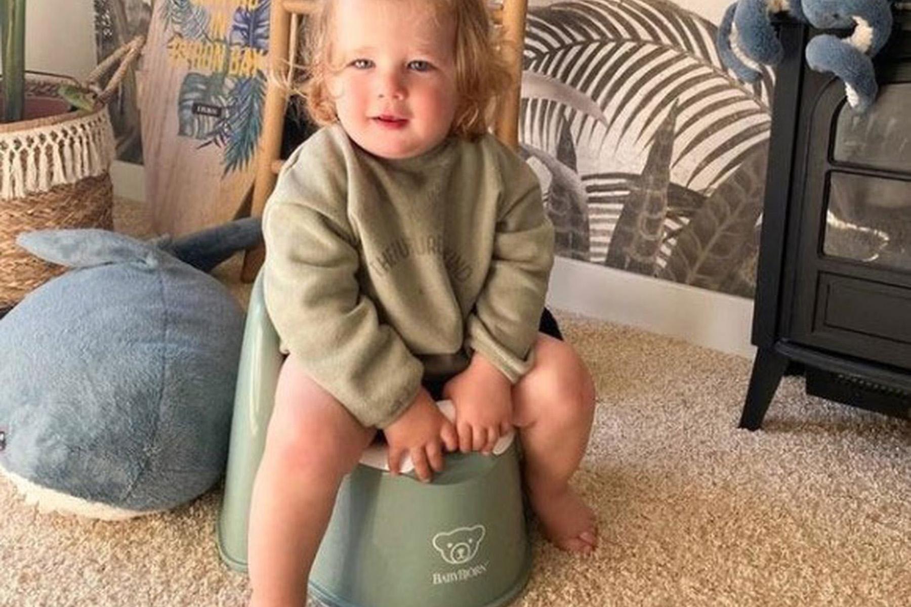 Toddler sitting on a light green potty, wearing a gray sweater, in a room with wallpaper and a plush toy on the floor.