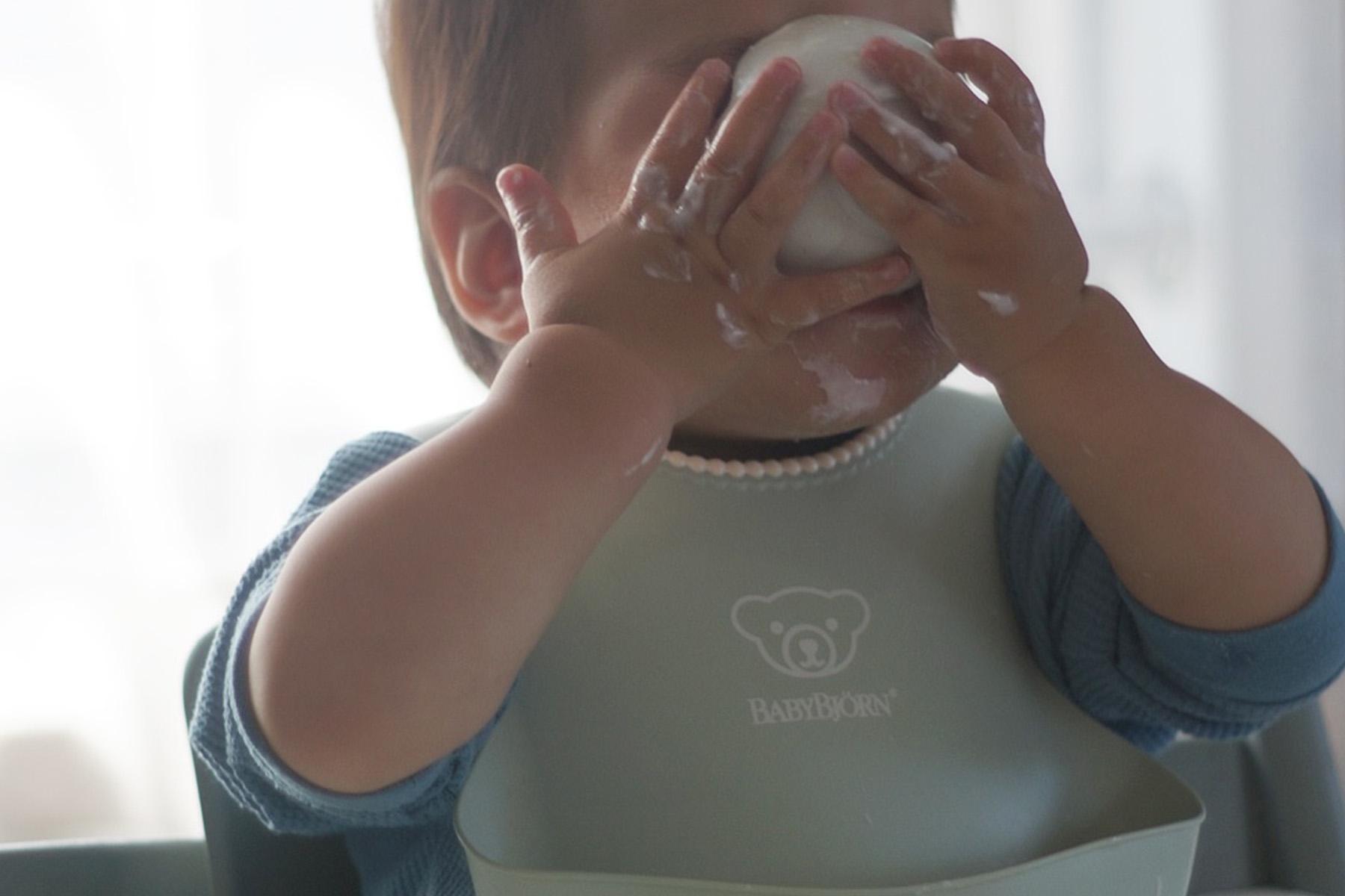 A toddler covering their face with a cup, wearing a green bib and a blue shirt, with food traces on their hands.