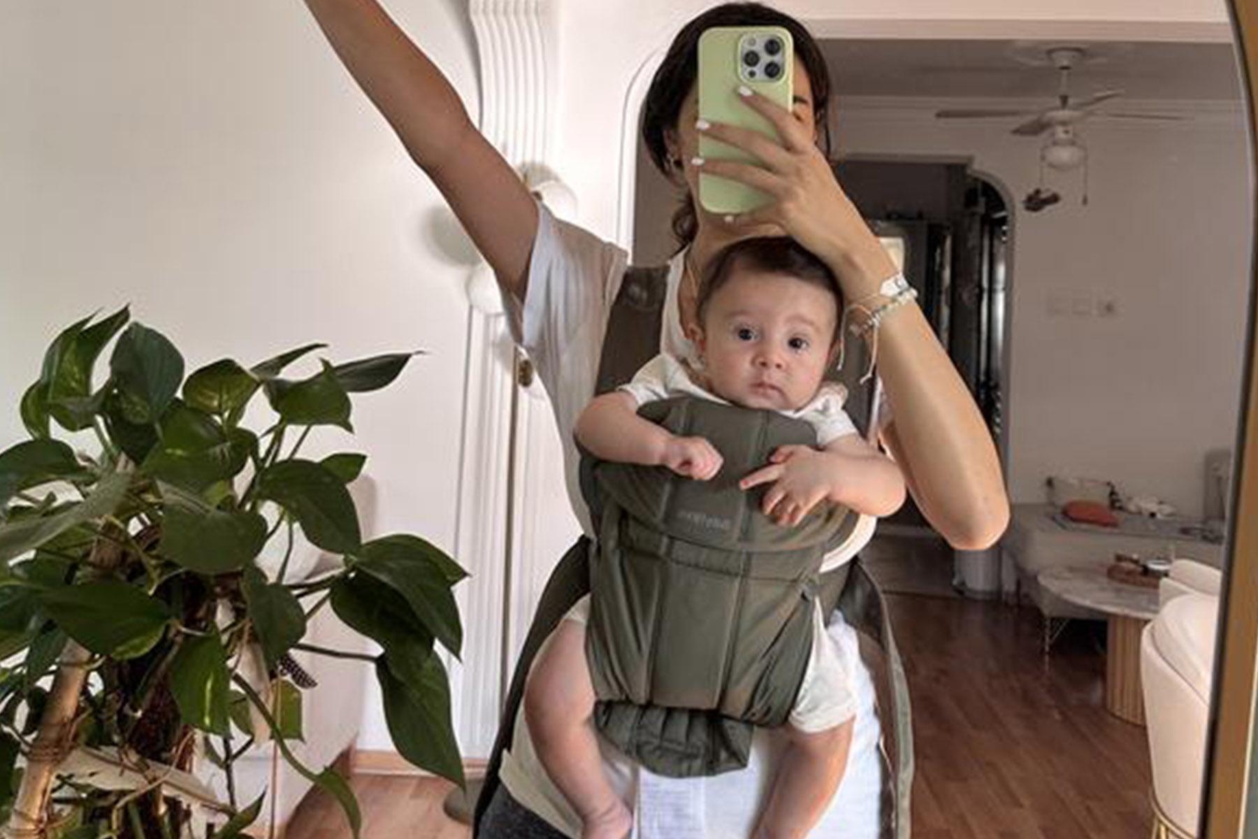 Mirror selfie of a person wearing a baby in an olive-green front carrier in a living room with a potted plant.