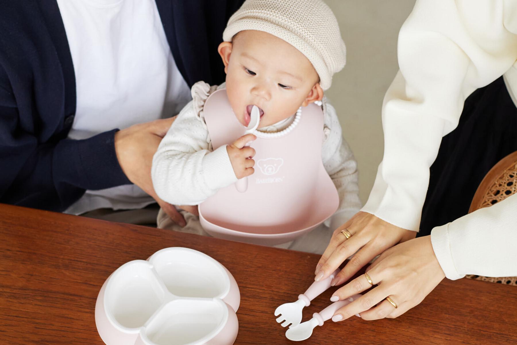 Baby wearing a bib and hat, sitting at a table, eating with a spoon. An adult's hands are arranging a divided plate nearby.