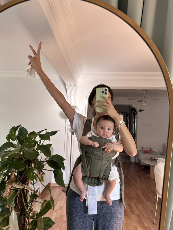 Person taking a mirror selfie with a baby in a green carrier, raising one arm in a peace sign. Room has wooden floors and a large plant.