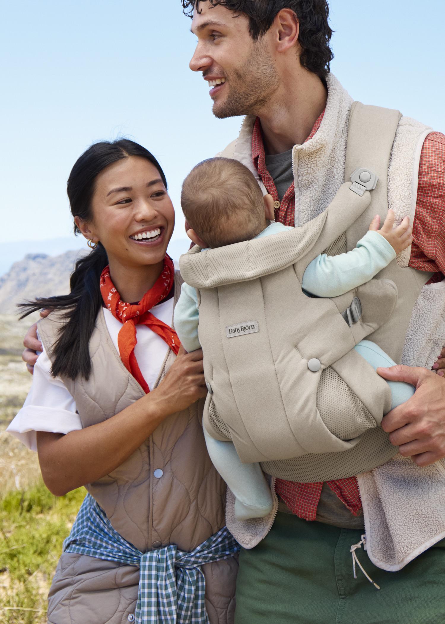 A smiling couple with a baby in a beige carrier, enjoying a sunny outdoor setting with mountainous scenery.