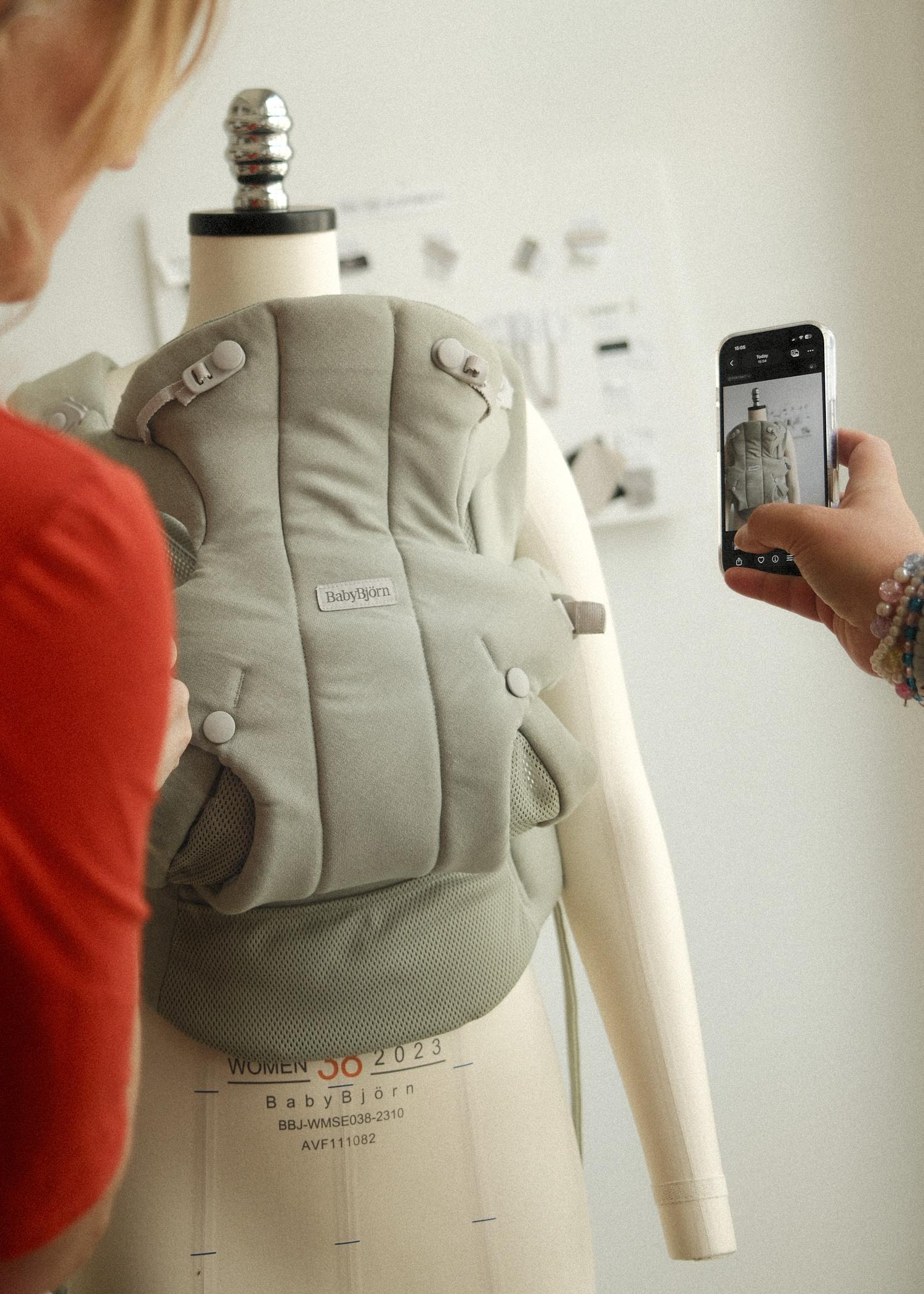 A woman photographs a beige baby carrier on a mannequin, labeled "BabyBjörn," with a blurred person on the side and a bulletin board in the background.