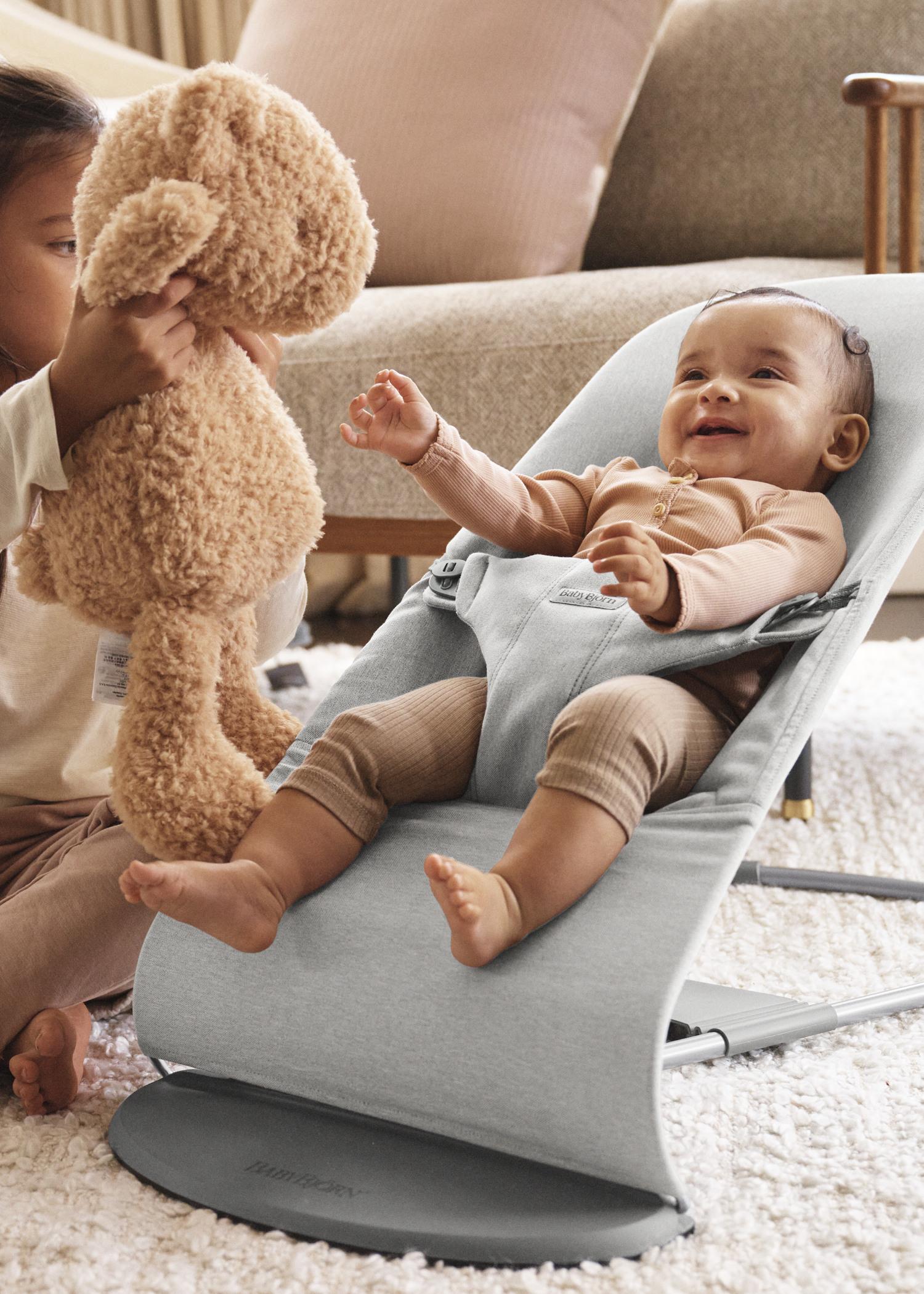 A baby in a bouncer smiles at a child holding a teddy bear, sitting on a soft carpet in a cozy living room.