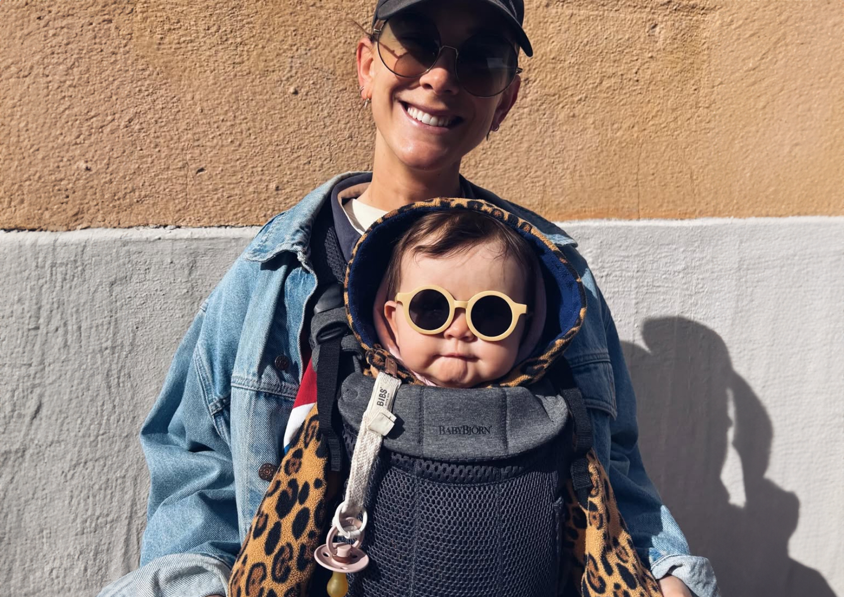 Smiling person in a cap and sunglasses carries a baby in a leopard print outfit and round sunglasses in a baby carrier.