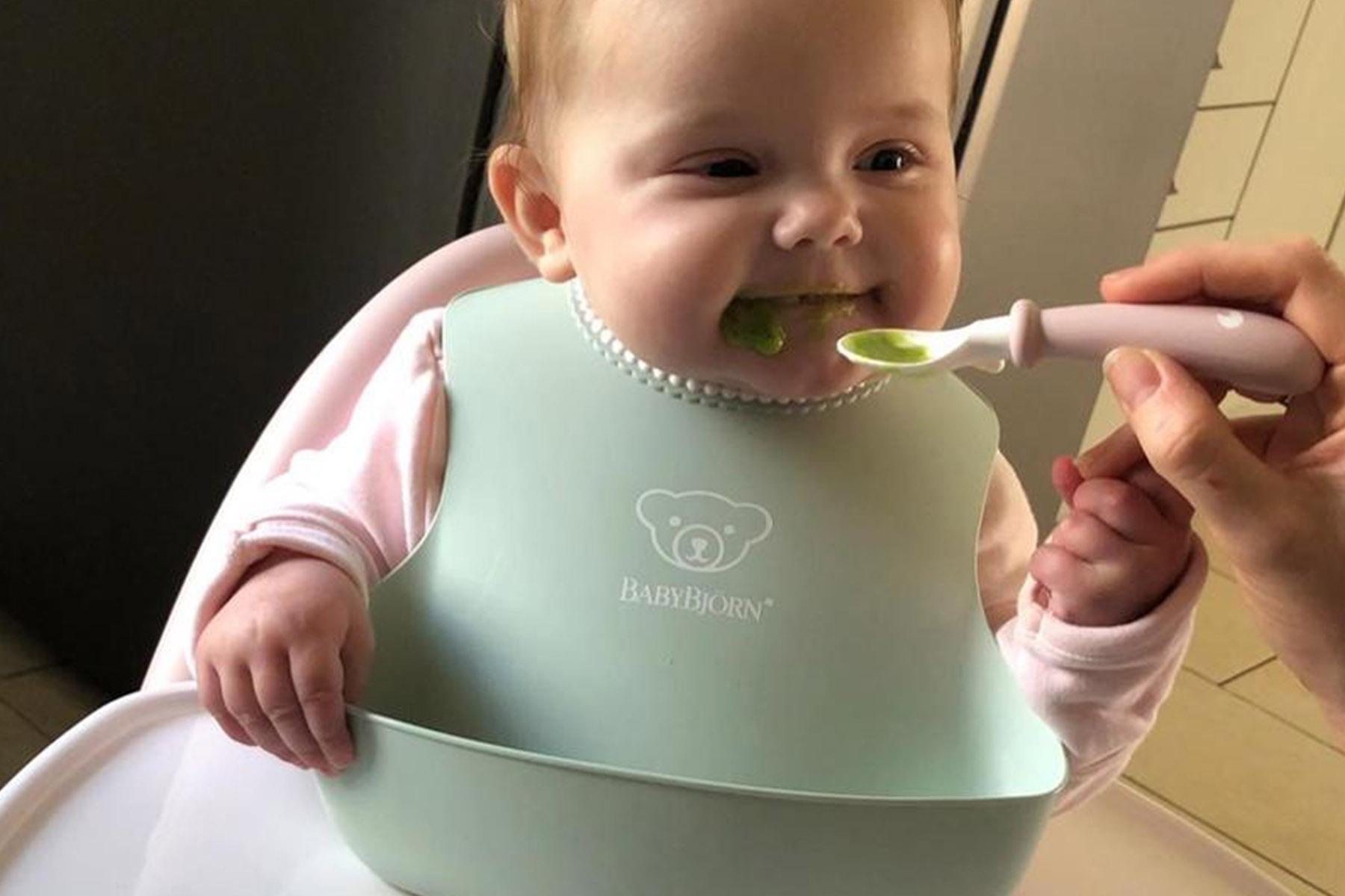 Smiling baby in high chair wearing mint-green bib being spoon-fed green puree.