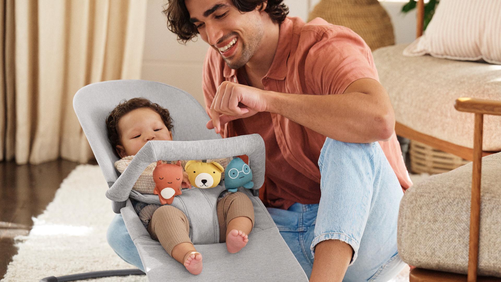 A man smiles at a baby sitting in a bouncer with plush toys, in a cozy living room with soft lighting and neutral decor.