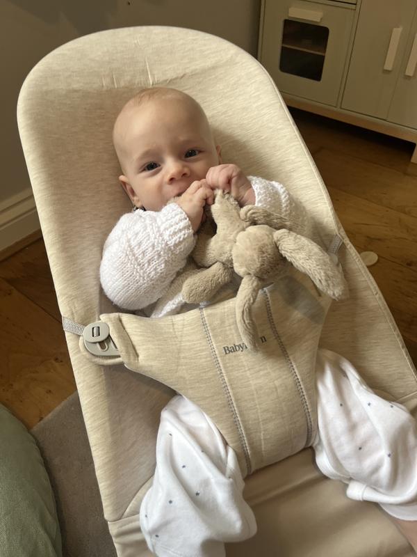 Baby in a beige bouncer, wearing a white outfit, holds a soft toy with both hands, sitting indoors on a wooden floor.