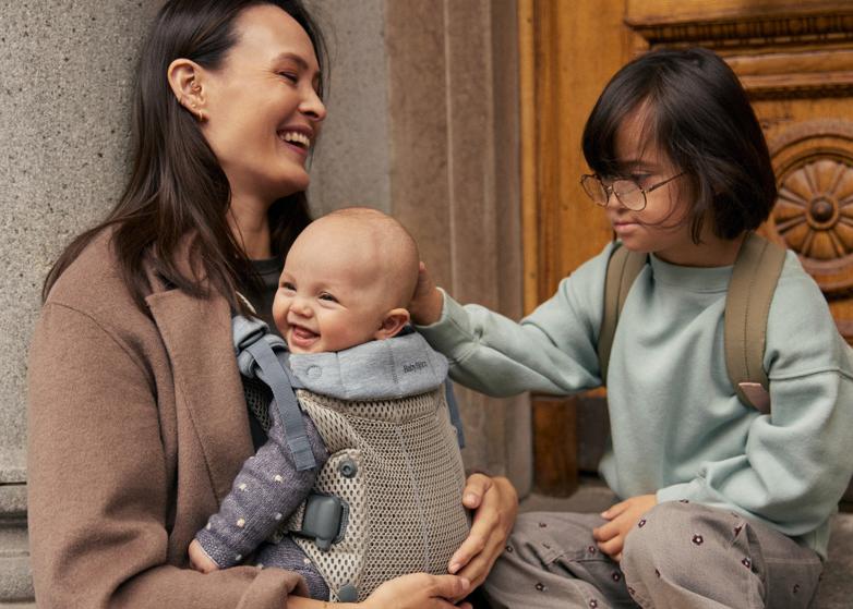A woman smiles while holding a baby in a carrier. A child next to them gently touches the baby's head. They sit by a stone pillar and wooden door.