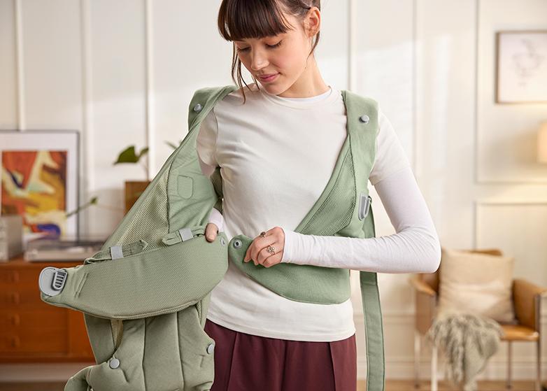 A woman is adjusting a green baby carrier with SnapWrap Design around her torso in a bright, cozy living room.